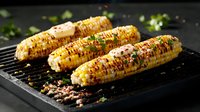 Grilled corn on the cob with melted butter, herbs and spices on black grill grate. Golden charred kernels garnished with fresh parsley and seasonings in dark setting.