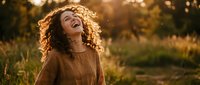Happy young woman with curly hair laughing joyfully in golden sunset light outdoors. Natural portrait expressing pure happiness and positive emotions in warm autumn setting.