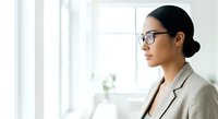 Young Asian woman wearing eyeglasses and beige blazer gazing out bright window in modern office setting, professional contemplation.