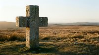 Stone cross monument standing alone in vast moorland landscape with golden grass and distant hills under soft natural light.