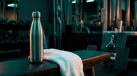 Stainless steel water bottle with towel on wooden bench in modern gym interior with teal lighting and equipment.