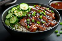 Glazed teriyaki pork slices served on steamed rice with fresh cucumber salad and green onions in black bowl, garnished with sesame seeds and sauce.