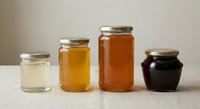 Four glass jars filled with different colored honey varieties from light to dark amber arranged on neutral background for organic food packaging.