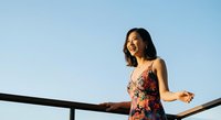 Young Asian woman in colorful floral dress laughing outdoors against clear blue sky, leaning on railing. Lifestyle and happiness concept for editorial and commercial use.