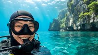 Young male scuba diver with mask and snorkel equipment swimming in crystal clear tropical waters near limestone cliffs covered in lush green vegetation.