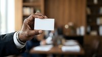 Man holding blank white business card in office environment with wooden interior and blurred background, suitable for corporate branding and professional networking concepts.