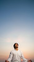 Man in white long-sleeved shirt meditating outdoors at sunset with arms outstretched, looking upward against clear sky. Suitable for wellness and mindfulness content.