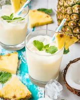 Fresh pineapple coconut smoothie garnished with mint leaves and fruit slices on turquoise background, summer tropical drink with ice cubes and paper straws.