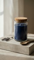 Glass jar with cork lid containing blue powder on concrete surface with wooden spoon and lavender sprigs in natural sunlight for organic skincare products.