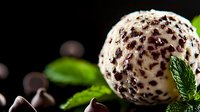 Creamy vanilla ice cream ball rolled in dark chocolate chips and mint leaves on black background, macro food photography for dessert menu or packaging.