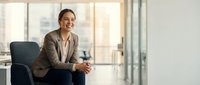 Professional businesswoman in beige suit sitting confidently in modern office chair, smiling at camera in bright corporate workspace for business content.