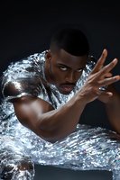 African American male model wrapped in reflective metallic material poses dramatically with outstretched hand against dark background, creating futuristic fashion portrait.