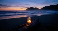 Glowing candle lantern on driftwood at twilight beach with purple sunset sky and ocean waves creating romantic coastal atmosphere for relaxation and meditation.