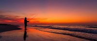 Silhouette of fisherman casting line on beach during golden sunset with dramatic sky colors reflecting on wet sand and ocean waves.