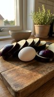 Fresh sliced eggplant and white egg on rustic wooden cutting board in bright kitchen with herbs and natural window light for cooking preparation.