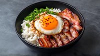 Asian rice bowl with crispy bacon, sunny side up egg, green onions and white rice in black ceramic bowl on dark background. Homemade comfort food.