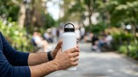 White insulated water bottle held by male hand in park setting with blurred people and green trees in background for beverage, lifestyle and outdoor advertising.
