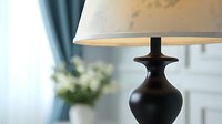 Elegant black ceramic table lamp with white shade casting warm light in modern interior setting with blue curtains and white flowers in background.
