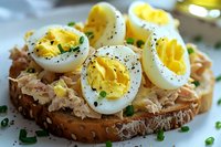 Tuna sandwich with sliced hard-boiled eggs on whole grain bread, garnished with fresh chives and black pepper, close up view of breakfast or lunch meal.