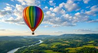 Colorful hot air balloon floating over scenic mountain landscape with winding river and green fields under dramatic cloudy sky at sunset.