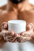 Male hands holding white moisturizer cream jar with water drops. Close up view of cosmetic product presentation for skincare and beauty industry.