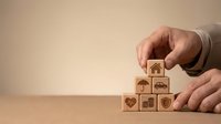 Businessman hands arranging wooden blocks with insurance icons including home, car, health and life protection symbols on neutral background.