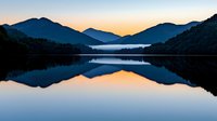 Tranquil mountain lake at dawn with perfect mirror reflection of silhouetted peaks and golden sunrise sky creating symmetrical landscape composition.