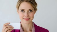 Young Caucasian woman with blonde hair in pink blazer holding blank business card against white background, smiling confidently at camera.