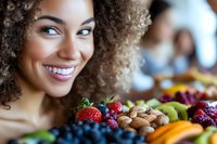 Young mixed-race woman smiling at colorful fresh fruit display with strawberries, blueberries, grapes and nuts at farmers market or grocery store.