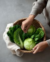 Female hands holding woven basket with fresh green napa cabbage and white cloth on gray background, top view. Organic vegetables for healthy eating.