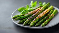 Grilled green asparagus spears seasoned with spices and fresh basil leaves served on gray ceramic plate against dark background. Fresh seasonal vegetables.