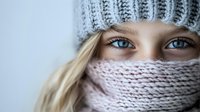 Close up portrait of young Caucasian woman with bright blue eyes wearing gray knitted winter hat and pink scarf wrapped around face against light background.