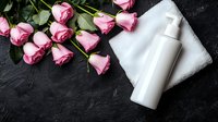 Pink roses and white cosmetic bottle on dark stone background with foam and towel, elegant spa and beauty care composition for skincare product presentation.