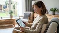 Middle-aged Caucasian woman in beige sweater using tablet computer while relaxing on gray sofa in bright modern living room with plants and natural light.