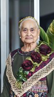 Senior Hispanic woman wearing traditional embroidered dress with lace details and golden headband, holding deep red roses against green foliage background.
