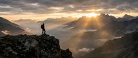 Lone hiker silhouette standing on rocky mountain peak at golden sunrise with dramatic sunbeams piercing through layered mountain ranges and misty valleys below.