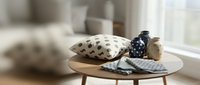 Cozy home decor arrangement with patterned pillows, decorative vase, and folded textiles on wooden table by window for interior styling and lifestyle content.
