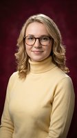 Professional blonde woman wearing glasses and yellow turtleneck sweater smiling confidently against burgundy background for business portraits.