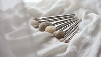 Professional makeup brushes with wooden handles arranged on soft white fabric for beauty and cosmetics applications.