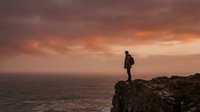 Silhouetted male hiker standing on rocky cliff overlooking vast ocean at sunset, wearing backpack. Dramatic golden and orange sky with clouds. Concept for adventure, travel, exploration, and outdoor lifestyle.