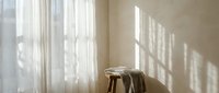 Minimalist interior with wooden stool and folded blanket by window with natural sunlight creating geometric shadows on neutral beige wall.
