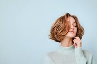 Young Caucasian woman with short wavy brown hair and closed eyes wearing light sweater shows natural beauty and serenity against blue background.
