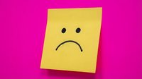 Yellow sticky note with sad face drawn in black ink on bright pink background expressing negative emotions and disappointment.