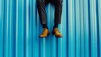 Young man legs in black pants and brown leather shoes sitting on blue corrugated metal wall background. Urban street style fashion concept for advertising.
