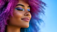 Young African American woman with vibrant purple curly hair and creative bright makeup smiling with eyes closed against blue background, expressing joy and confidence.