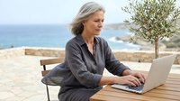 Mature woman with gray hair and light complexion, wearing dark casual shirt, working on laptop at wooden table with olive tree and coastal seascape backdrop, remote work and digital nomad lifestyle.