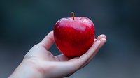 Fresh red apple held gently in female palm against dark blurred background. Clean minimalist composition highlighting healthy eating and natural food choices.
