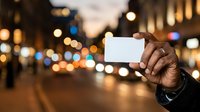 Young adult male with dark skin tone holding blank white card in urban evening street setting with blurred city lights and buildings for advertising or promotional messaging.