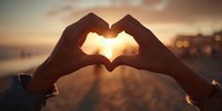 Hands forming heart shape silhouette against golden sunset over ocean water. Romantic love symbol gesture at beach during warm evening light for Valentine's Day concepts.