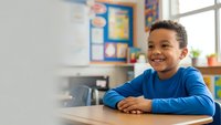 Happy African American elementary school boy smiling at desk in bright colorful classroom with educational posters and learning materials on walls.